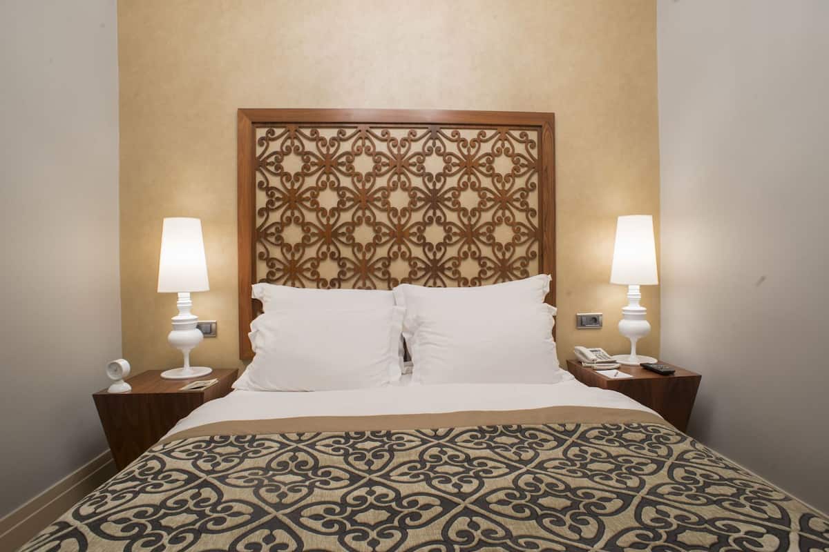 A hotel room with a patterned bedspread, two bedside lamps, a wooden headboard with a decorative pattern, and a telephone on the nightstand.
