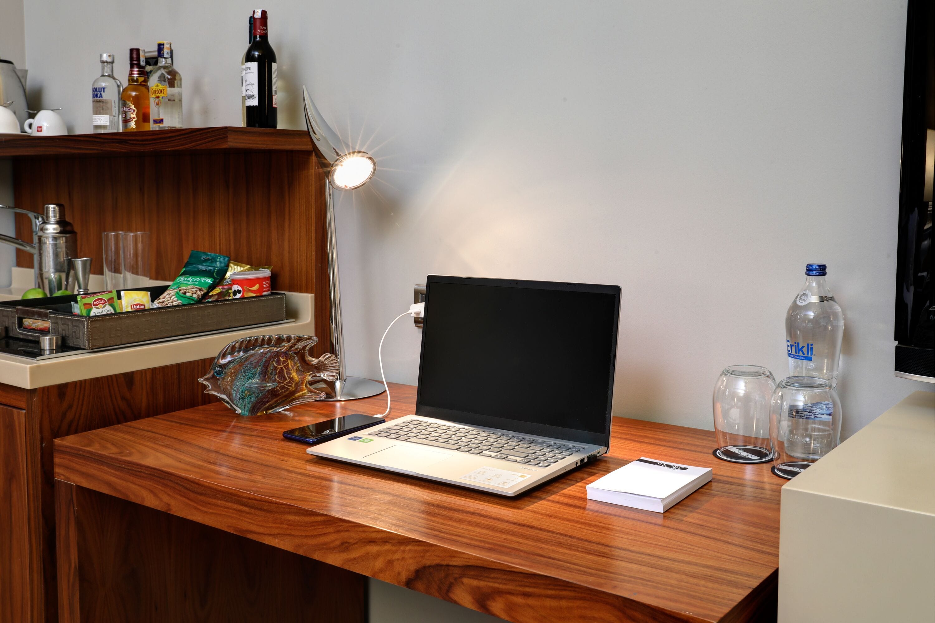 A wooden desk with a laptop, a smartphone, and a lamp. There are bottles and a glass on the desk.