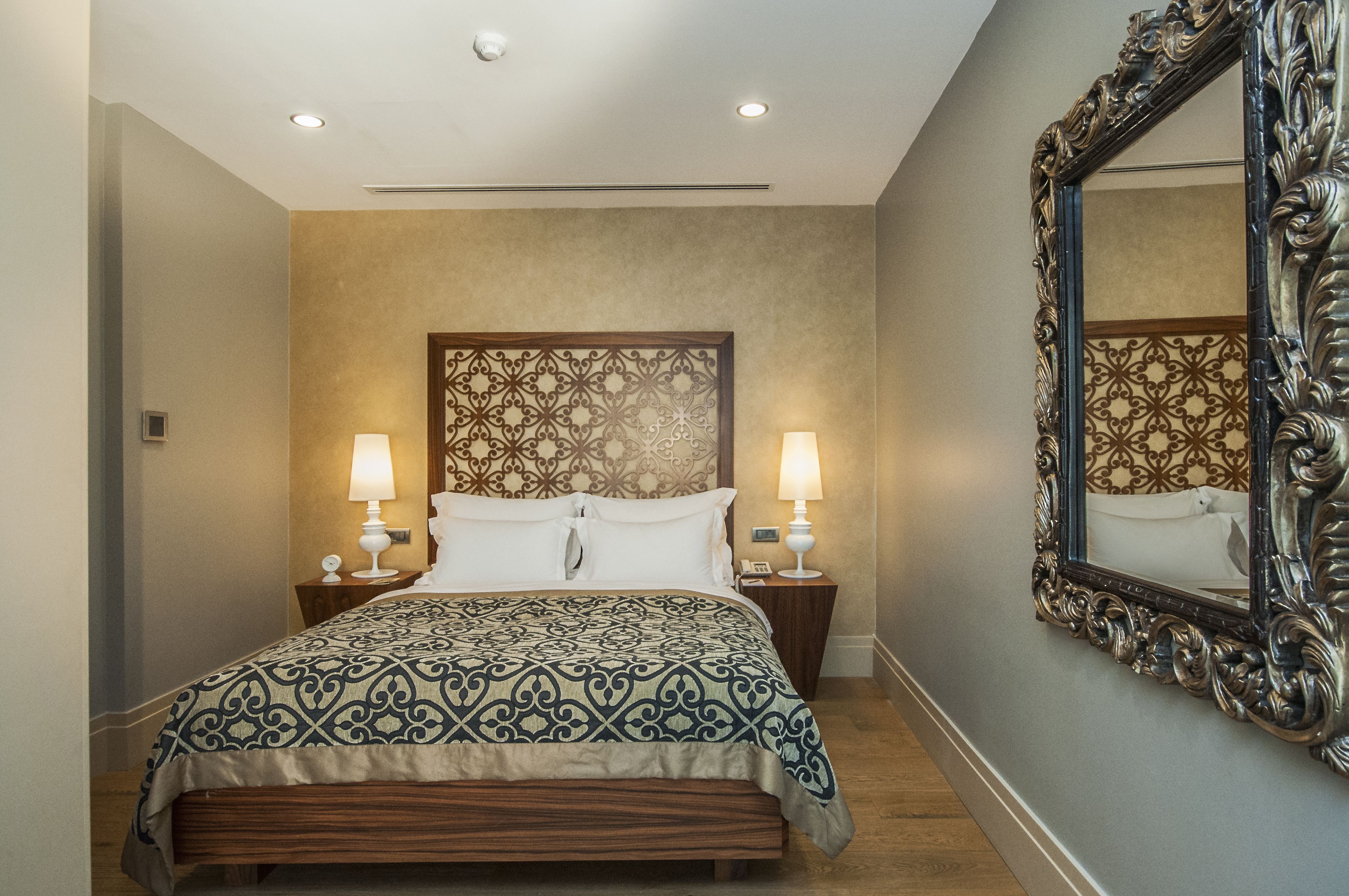 A hotel room with a large bed, two bedside lamps, a decorative headboard, and a large mirror with an ornate frame.