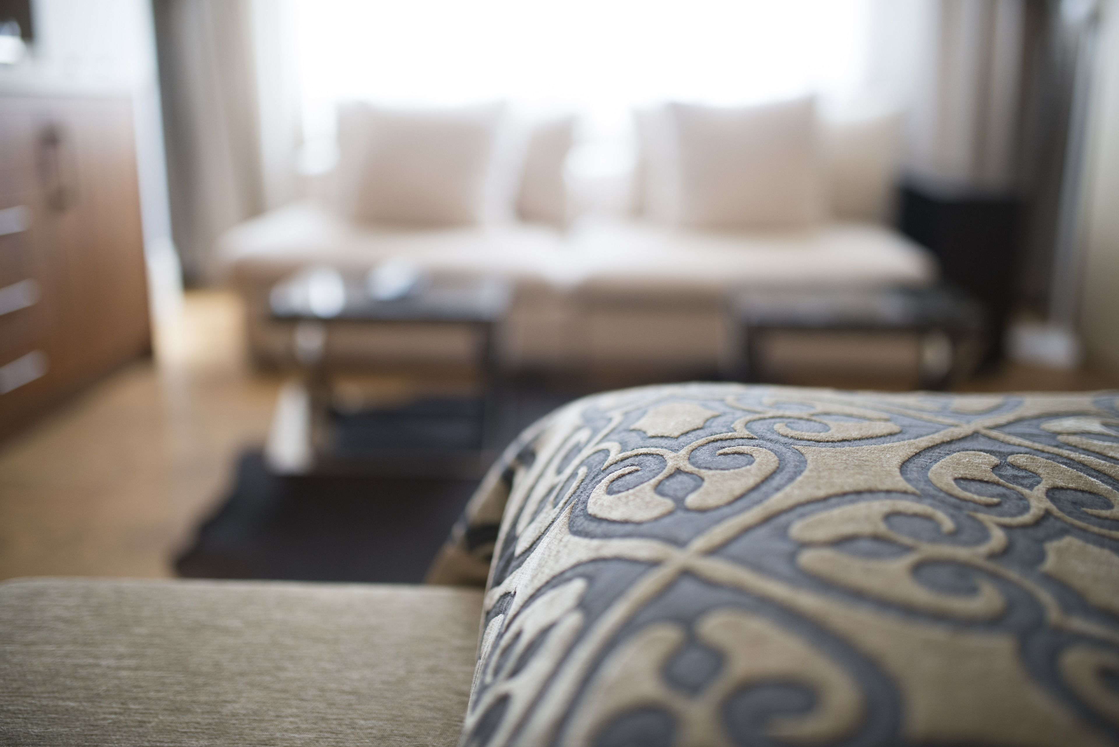 A close-up of a patterned bedspread with a blurred background featuring a bed and a dresser.