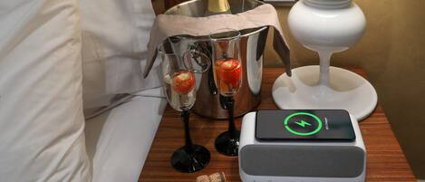 A hotel room with a bed, a bedside table with a bottle of champagne, two glasses with strawberries, a candle, and a power bank.