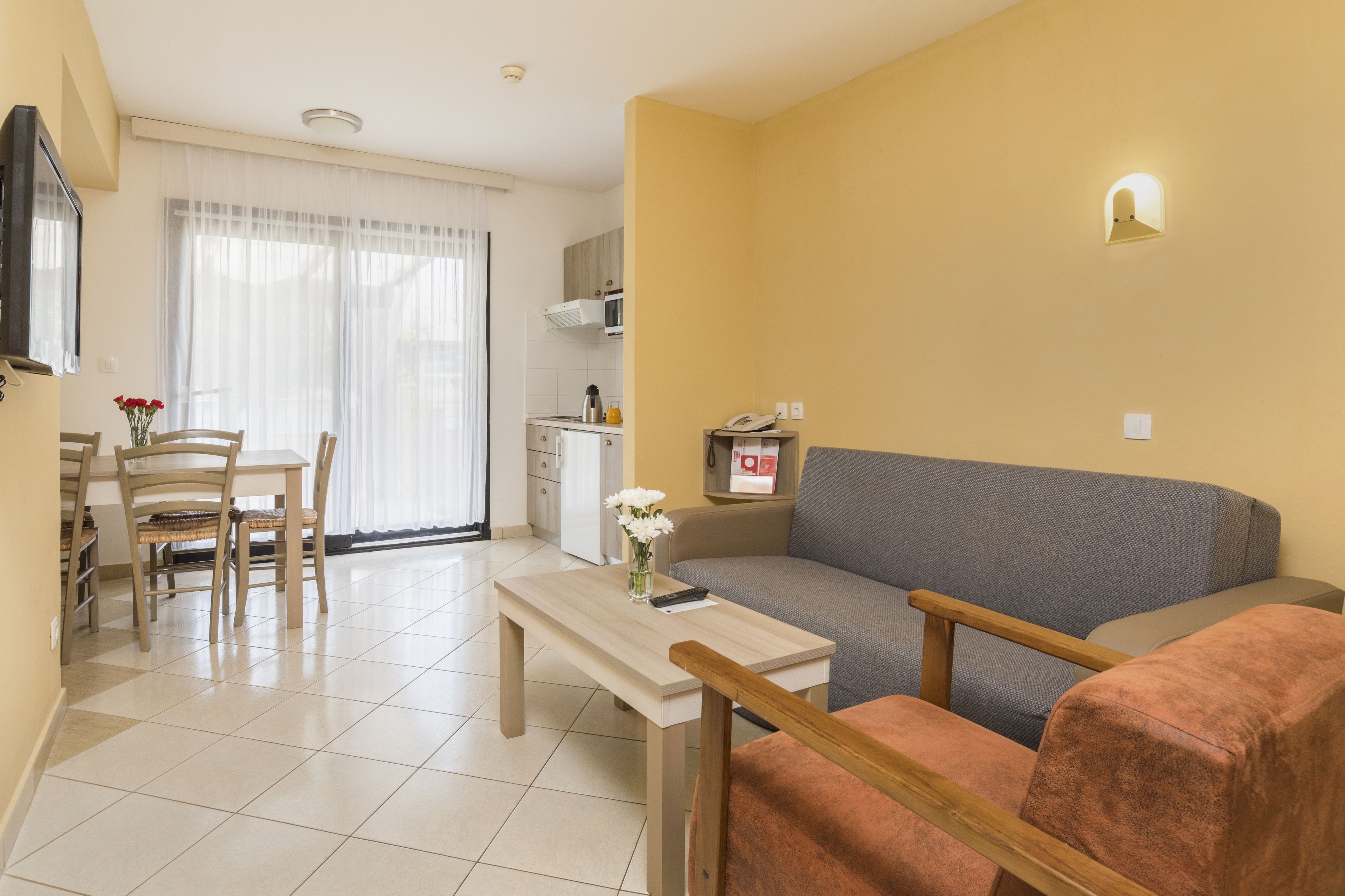 Ein Wohnzimmer mit einer Couch, einem Couchtisch und zwei Sesseln. Es gibt einen Esstisch mit Stühlen und ein Fenster mit Vorhängen.