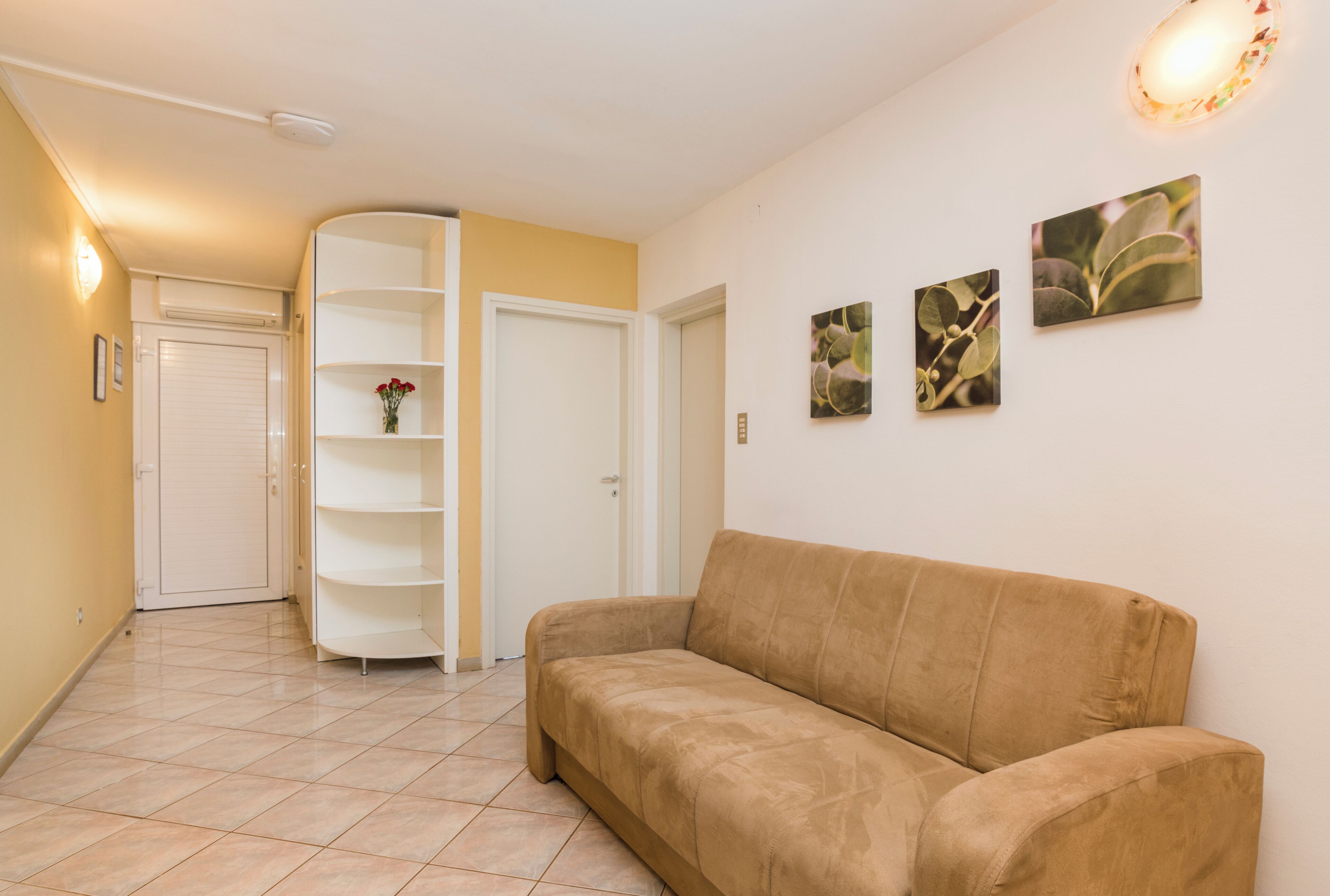Un salon avec un canapé beige, une étagère blanche et une porte donnant sur une autre pièce.