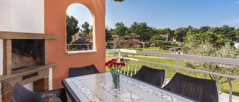 Une terrasse extérieure avec une cheminée en briques, une table ornée d’un vase de fleurs rouges, des chaises en osier et une vue sur un quartier résidentiel avec des maisons et des arbres.