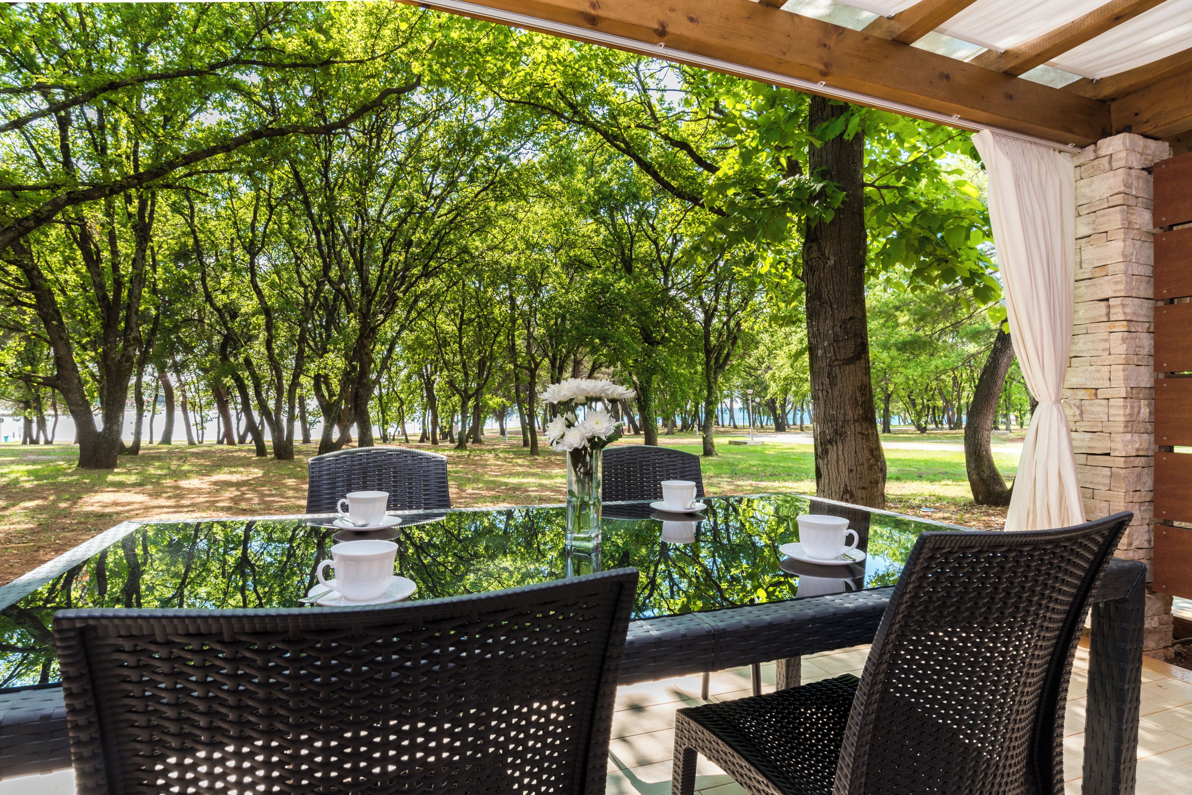 Un espace de restauration en plein air, avec des chaises en osier et une table en verre, entouré d’arbres et d’un mur de briques.