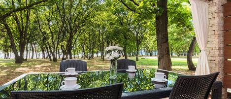 An outdoor dining area with wicker chairs and a glass table, surrounded by trees and a brick wall.