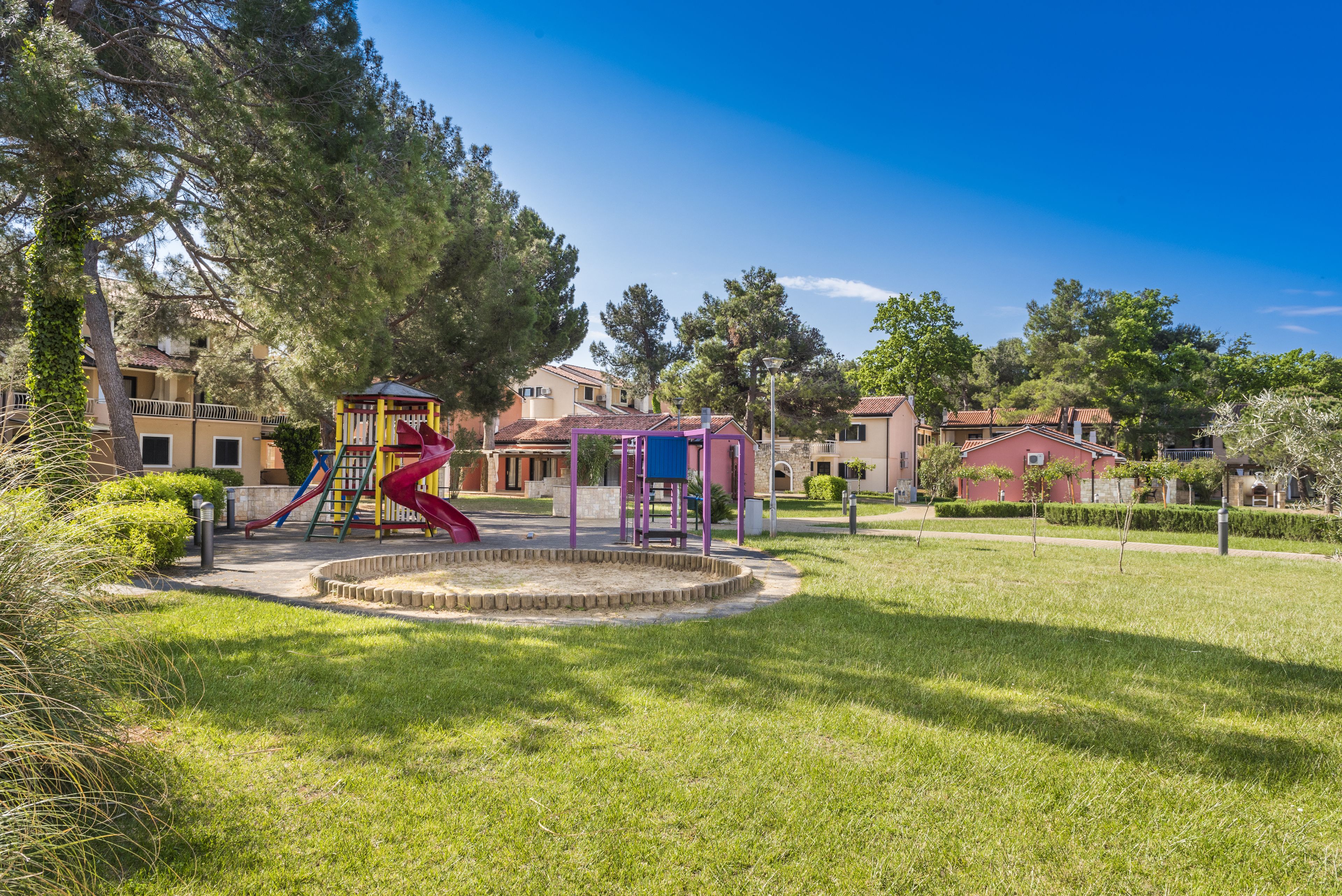A playground with slides and a climbing structure, surrounded by a well-maintained lawn and trees.