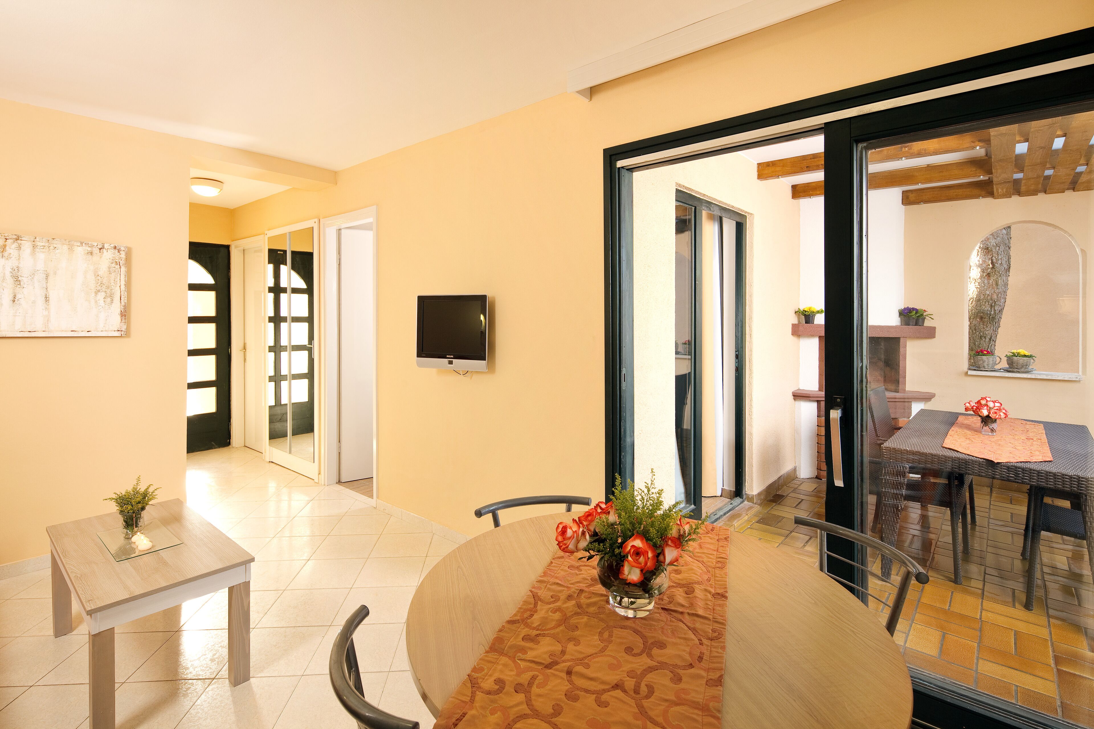 A dining area with a round table, chairs, a vase with flowers, a television, and a sliding glass door leading to an outdoor patio.