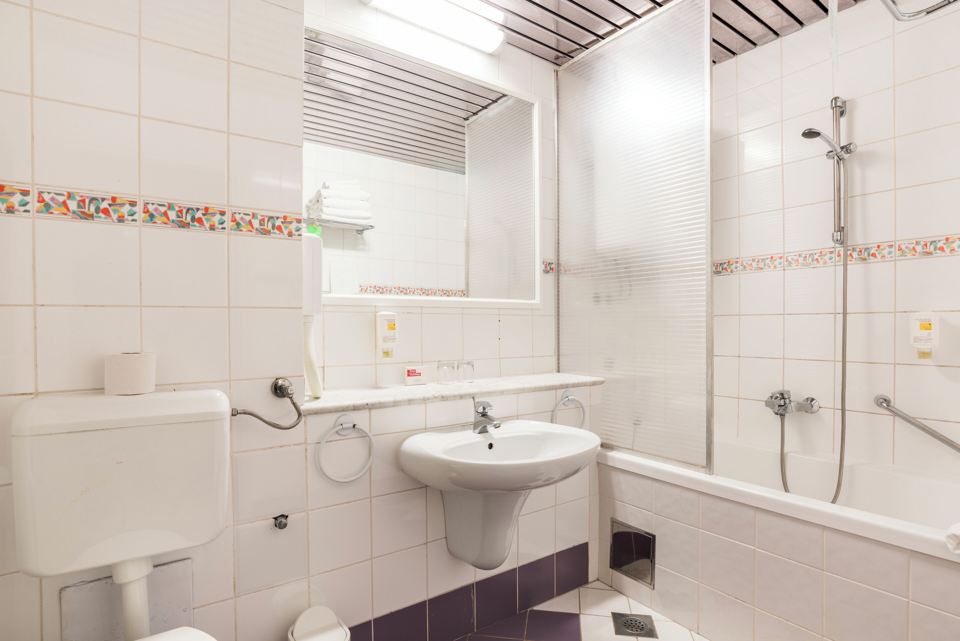 Une salle de bain équipée de toilettes, d’un lavabo, d’un miroir, d’une douche et de murs carrelés.