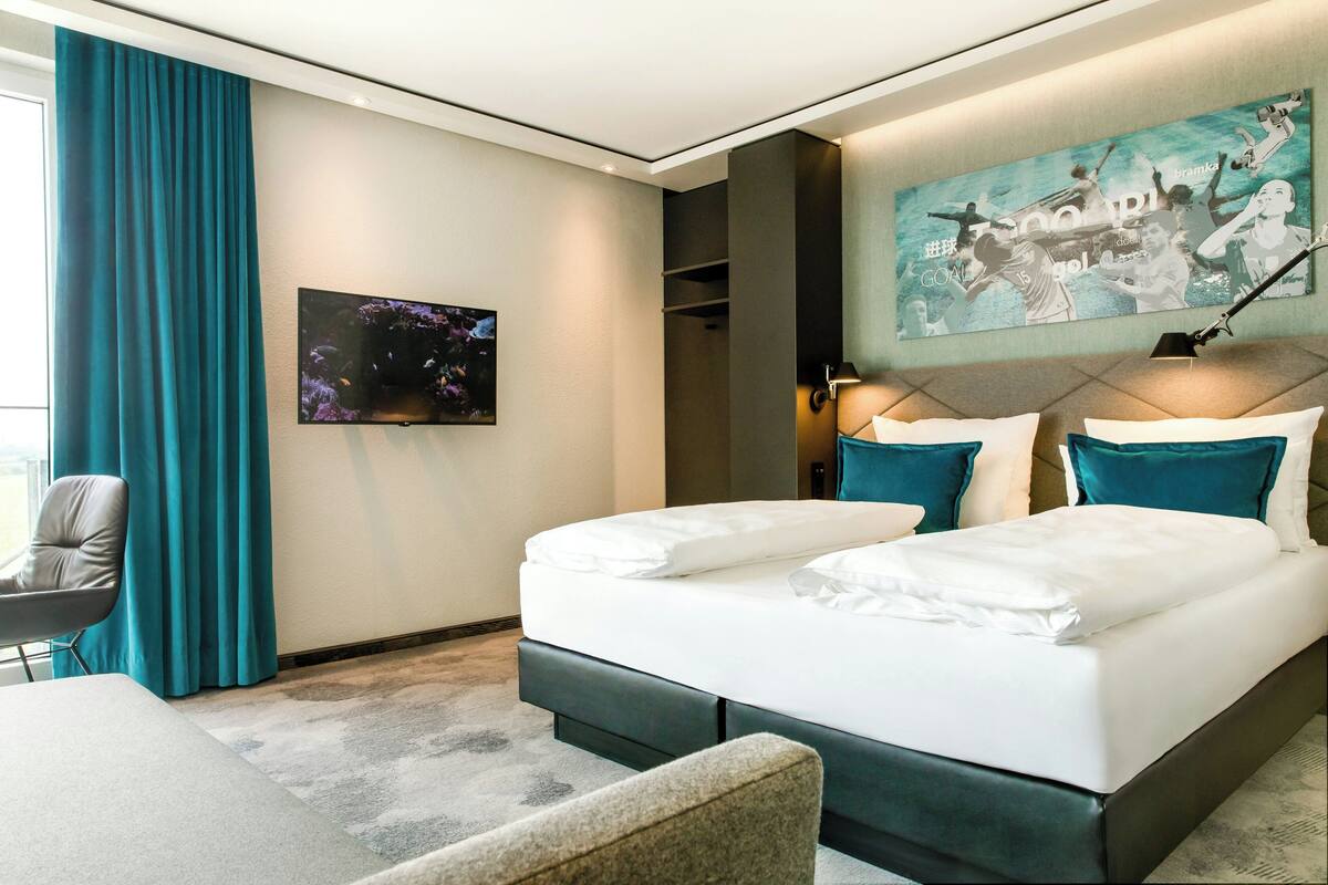 A modern hotel room with a large bed, a chair, a desk, and a painting on the wall.