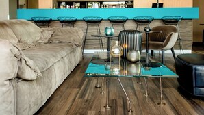 A modern living room with a sofa, a glass coffee table, and a bar area with chairs and a shelf of bottles. - Motel One München-Garching (Garching)
