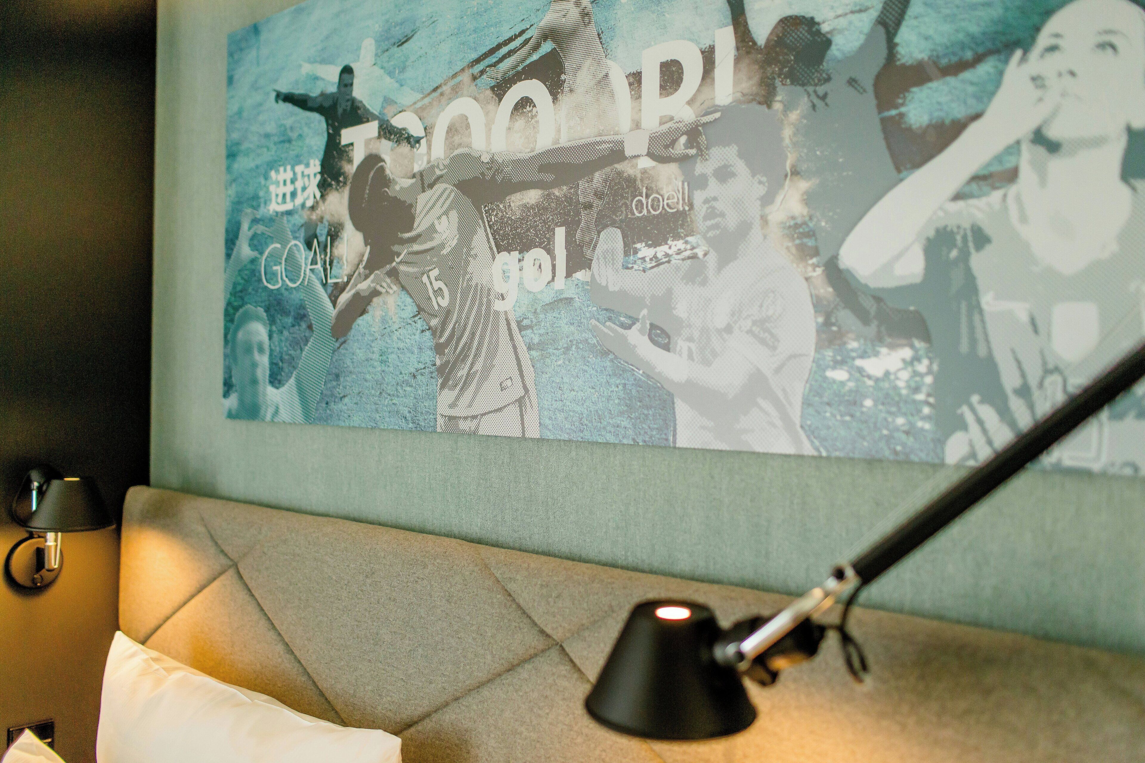 Een muur met sportkunst, een bed met een kussen en een bureaulamp.