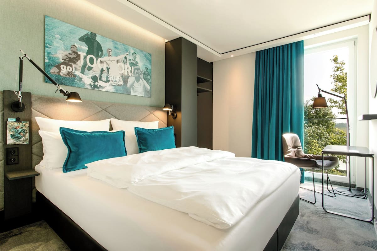 A modern hotel room with a large bed, a desk, and a view of the outdoors.