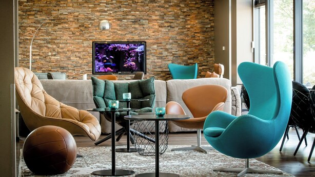 A modern living room with a brick wall, comfortable seating, a television, and a large window. - Motel One München-Garching (Garching)