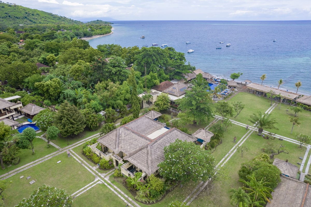 Vue aérienne d’un complexe hôtelier avec une piscine, un grand bâtiment doté d’un toit en tuiles et une plage où se trouvent des bateaux.