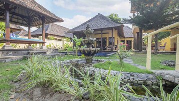 Un patio con una fuente de piedra, construcciones con techo de paja y una piscina.