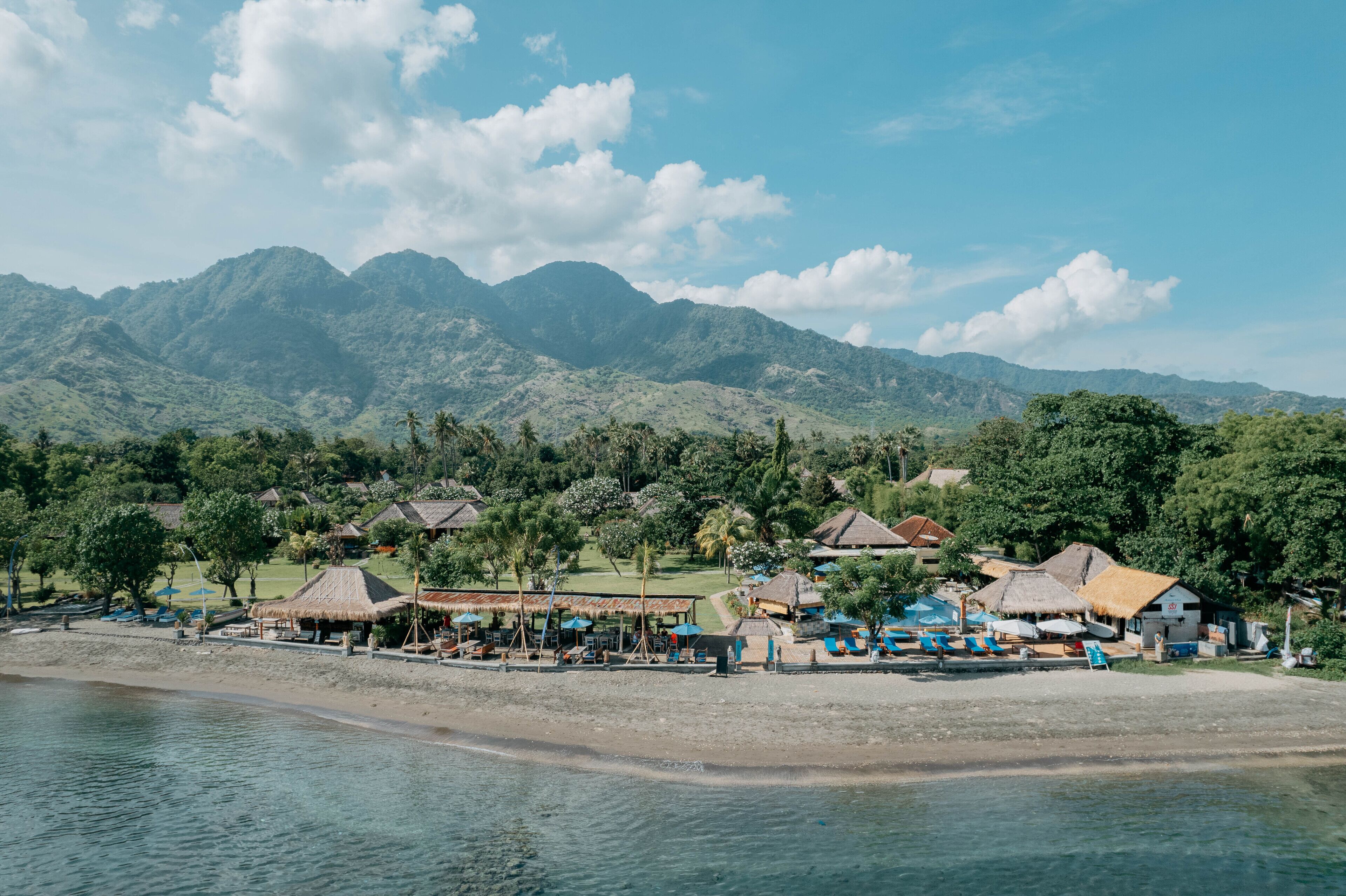 A beachside resort with thatched-roof huts, a pool area, and a view of mountains.