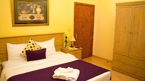 A neatly made bed with a purple bedspread and white linens, a wooden nightstand with a lamp and a telephone, a wooden wardrobe, and a framed picture on the wall.