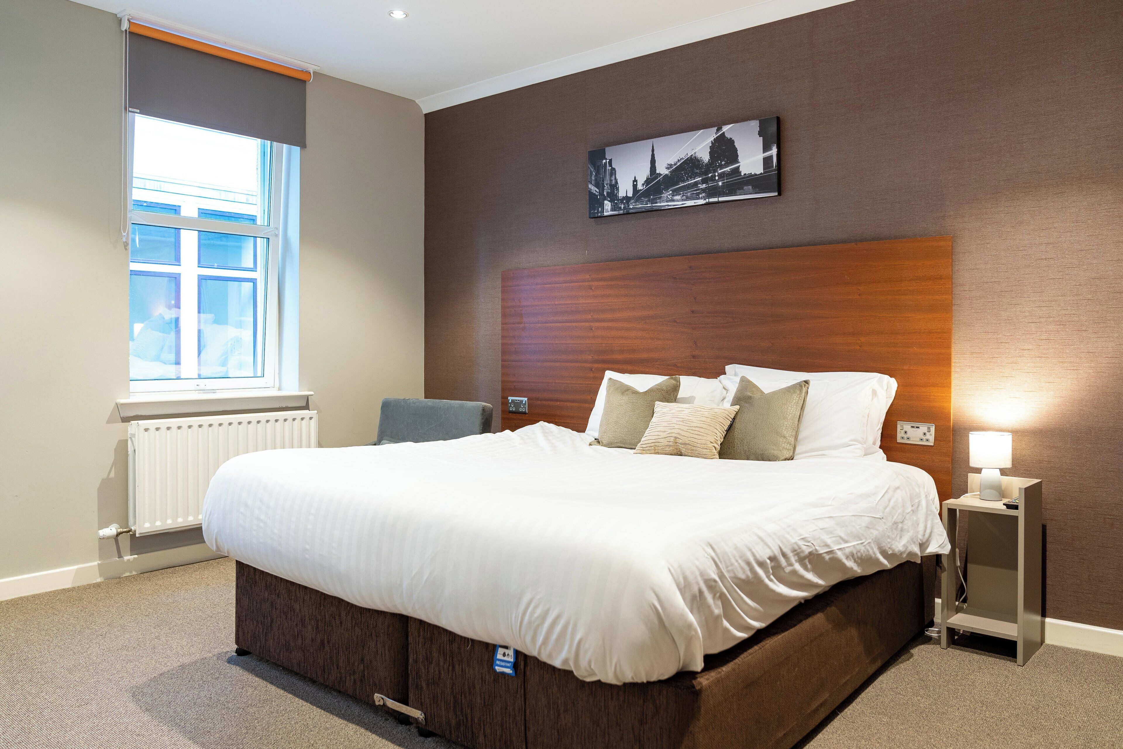A neatly made bed with a wooden headboard, a bedside table with a lamp, and a window with blinds.