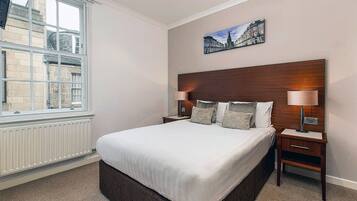 A hotel room with a large bed, two bedside tables, a headboard, a window with a view of buildings, and a painting on the wall.