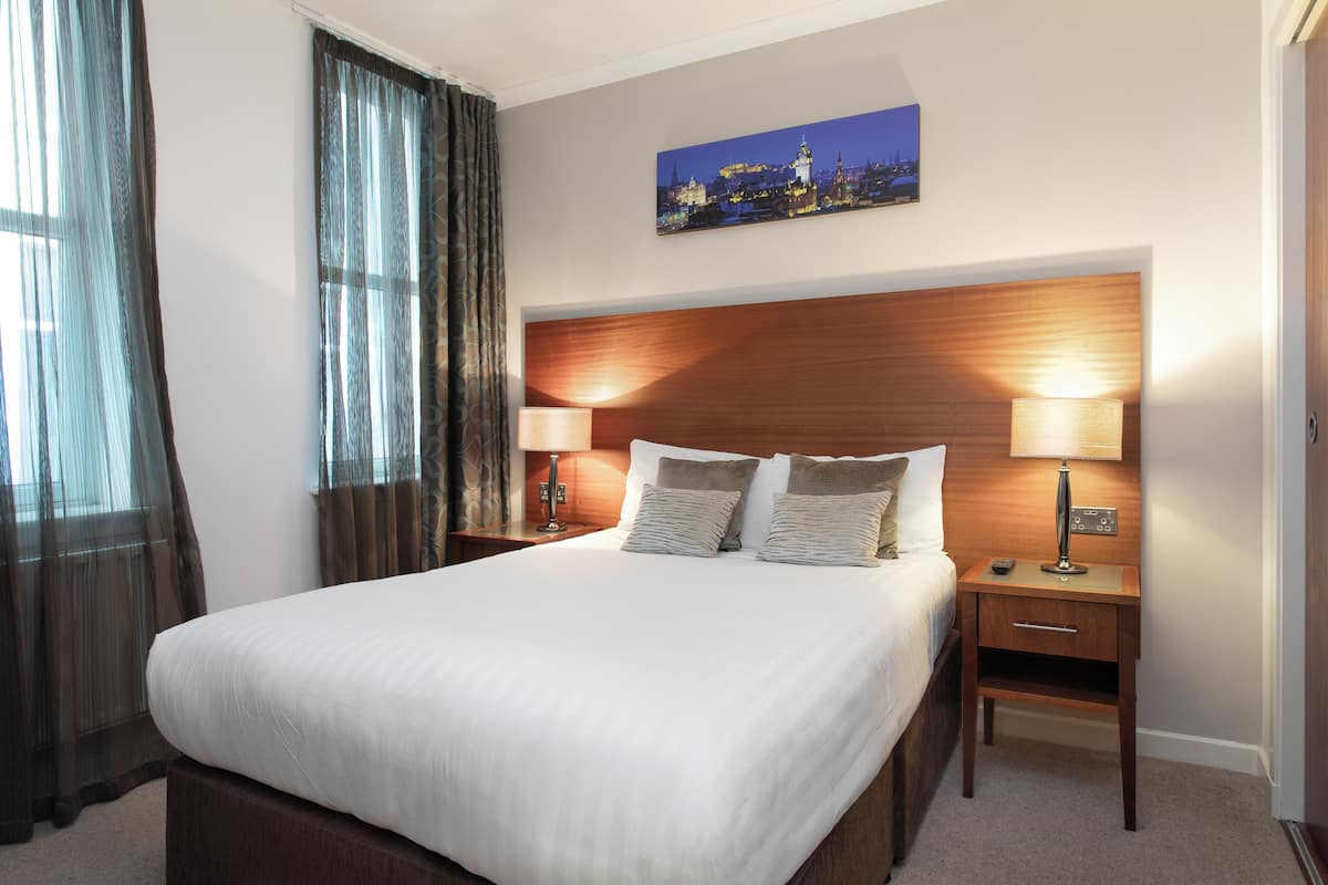 A hotel room with a large bed, two bedside tables with lamps, a headboard, and a cityscape painting on the wall.