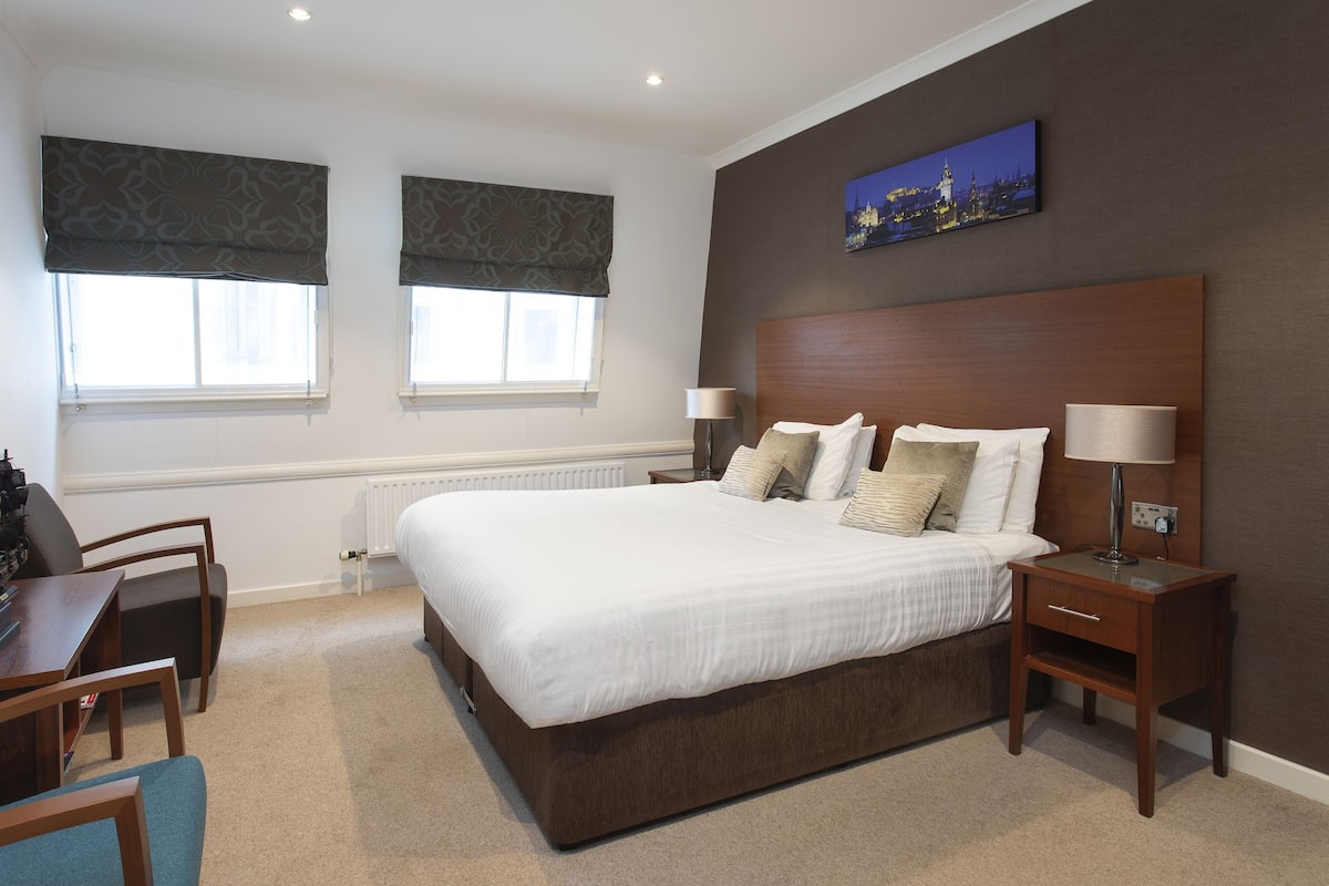 A hotel room with a large bed, two bedside tables, a desk with a chair, and two windows with curtains.