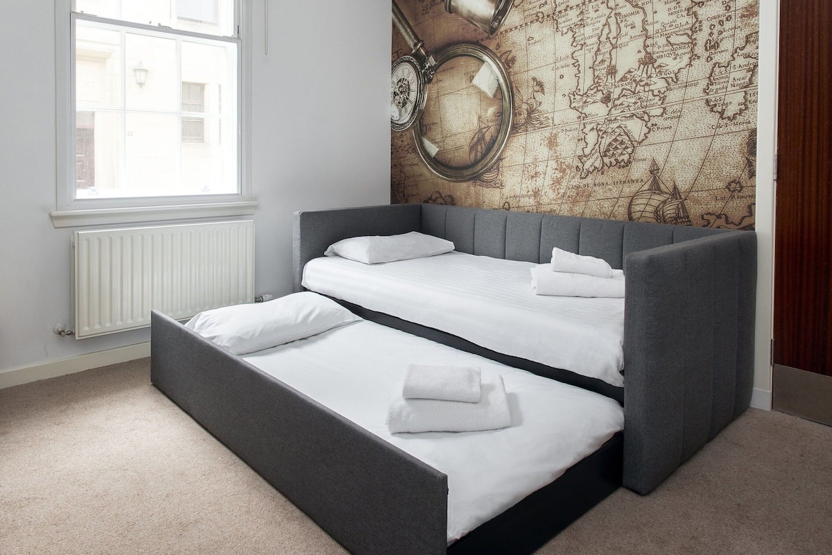 A room with a bed featuring a grey headboard and white bedding, a large window with a view of buildings, and a wall with a vintage map design.