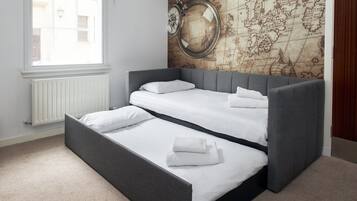 Una habitación con cama, cabecera gris y ropa de cama blanca, un ventanal con vistas a edificios, y una pared decorada con un diseño de mapa antiguo.