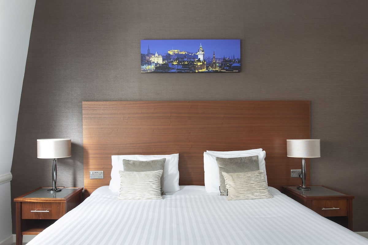 A hotel room with a large bed, two bedside tables with lamps, a nightstand, and a cityscape painting on the wall.
