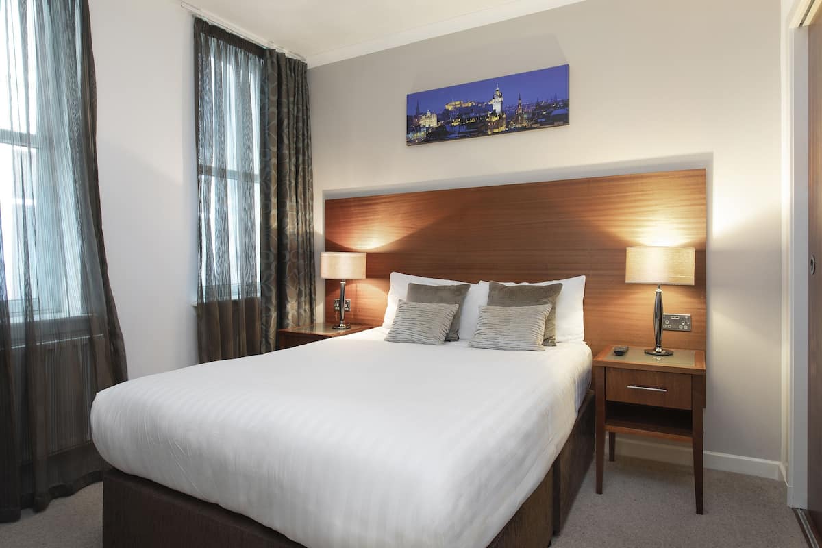 A hotel room with a large bed, two bedside tables with lamps, a cityscape painting on the wall, and sheer curtains on the window.