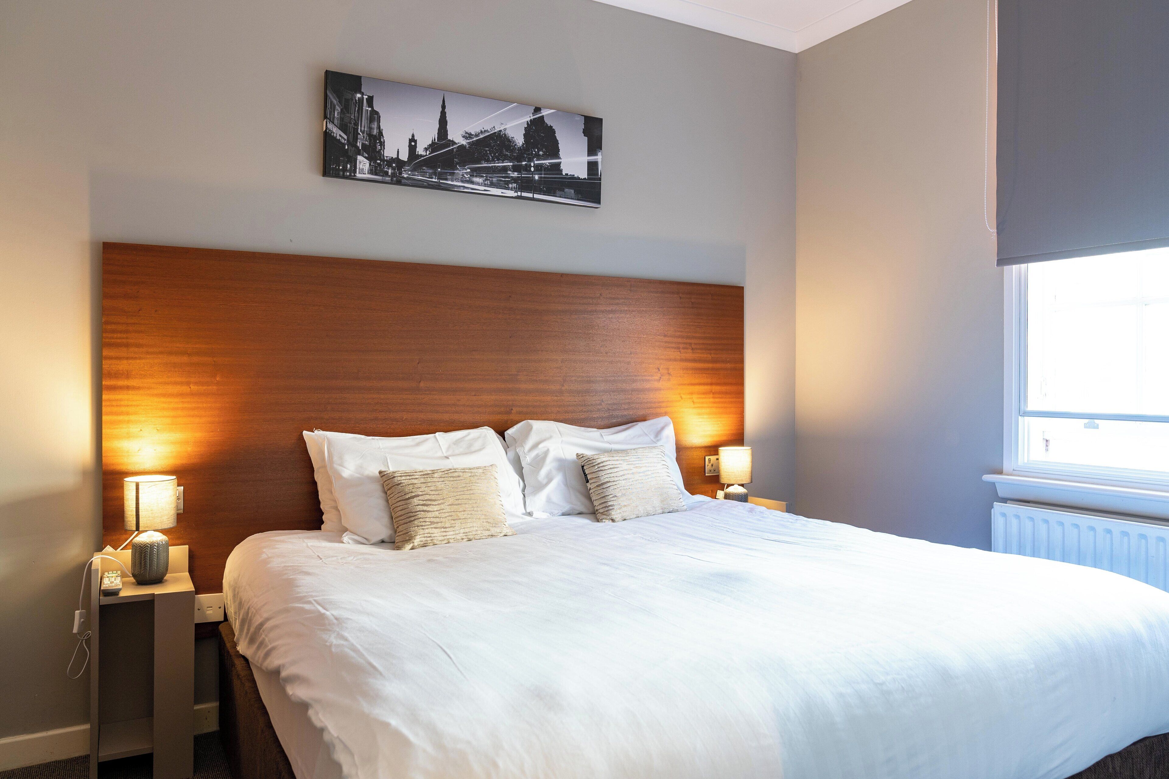 A hotel room with a large bed, wooden headboard, two bedside tables with lamps, a framed picture on the wall, and a window with daylight.