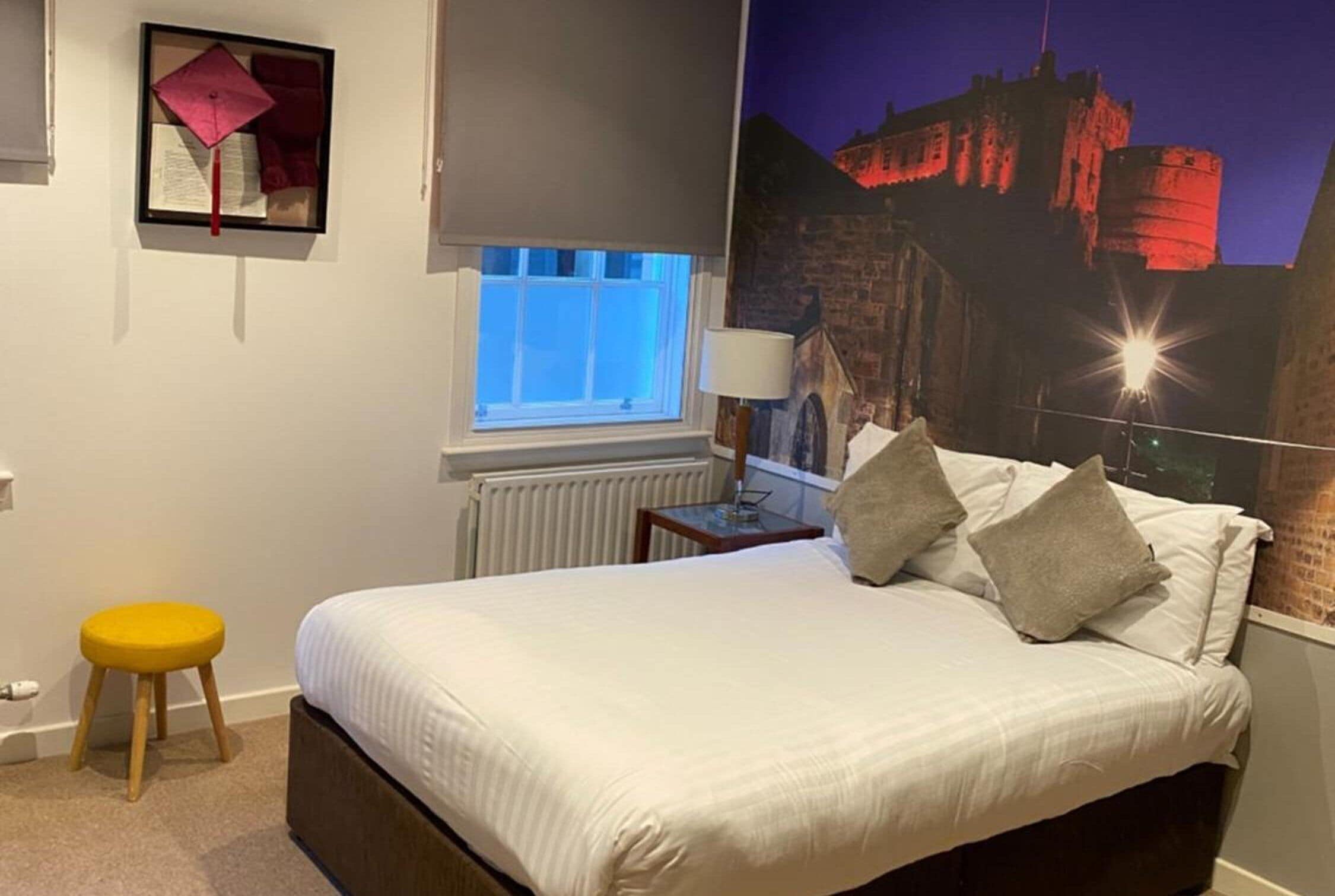 A bedroom with a large bed, a nightstand, a window, and a wall-mounted artwork of a castle.