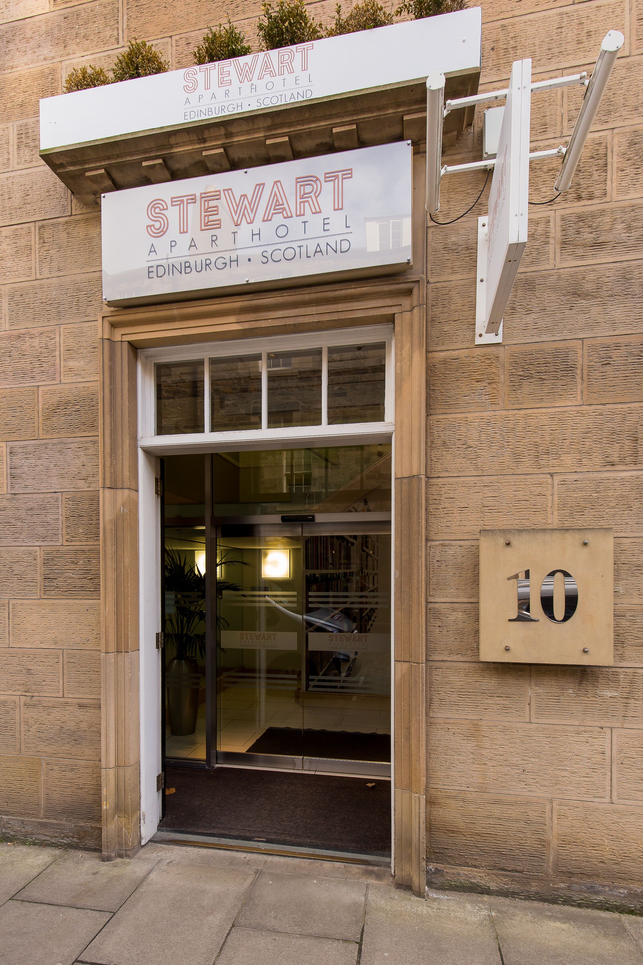 The exterior of the Stewart APT Hotel in Edinburgh, Scotland, featuring a large glass entrance, a signboard above the entrance, and a number plate indicating the address.