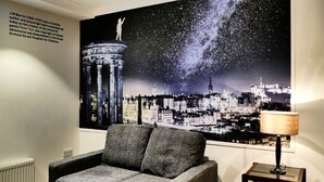 A room with a grey sofa, a lamp, and a large poster of a cityscape with stars. - Stewart by Heeton Concept (Edinburgh)