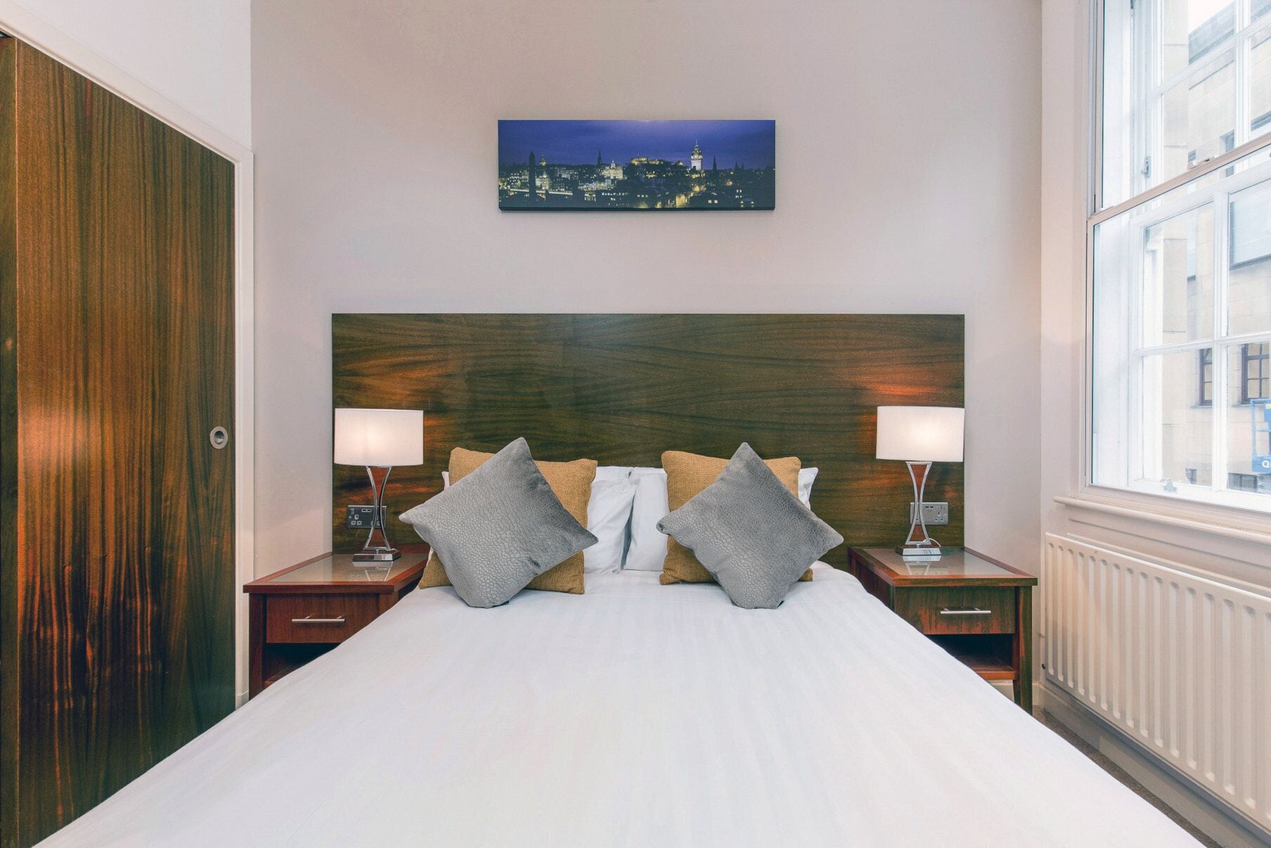 A hotel room with a large bed, two nightstands with lamps, a cityscape painting on the wall, and a window with a view of buildings.