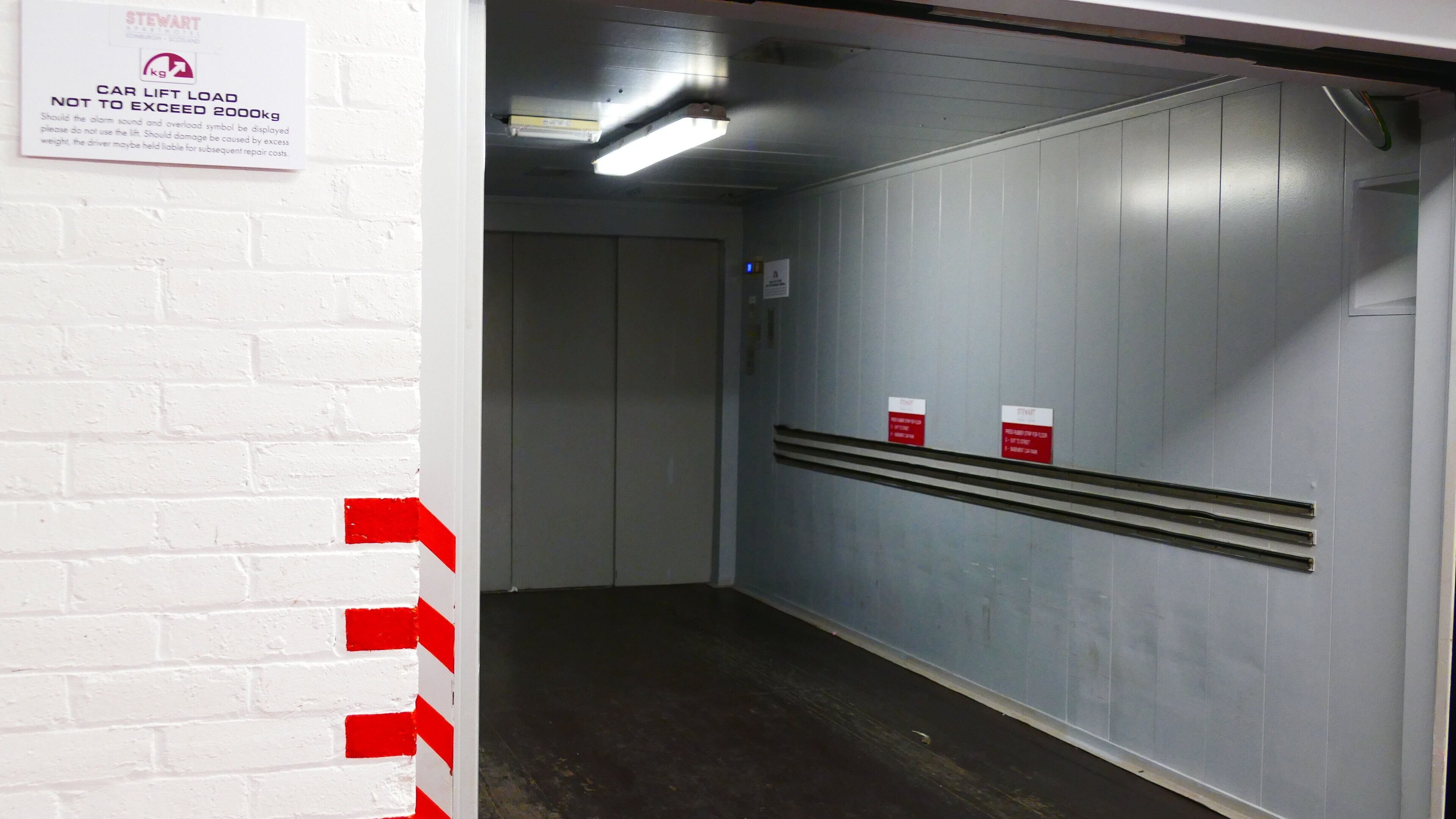 A car lift area with a warning sign, a white brick wall, and a grey wall with ventilation grilles.