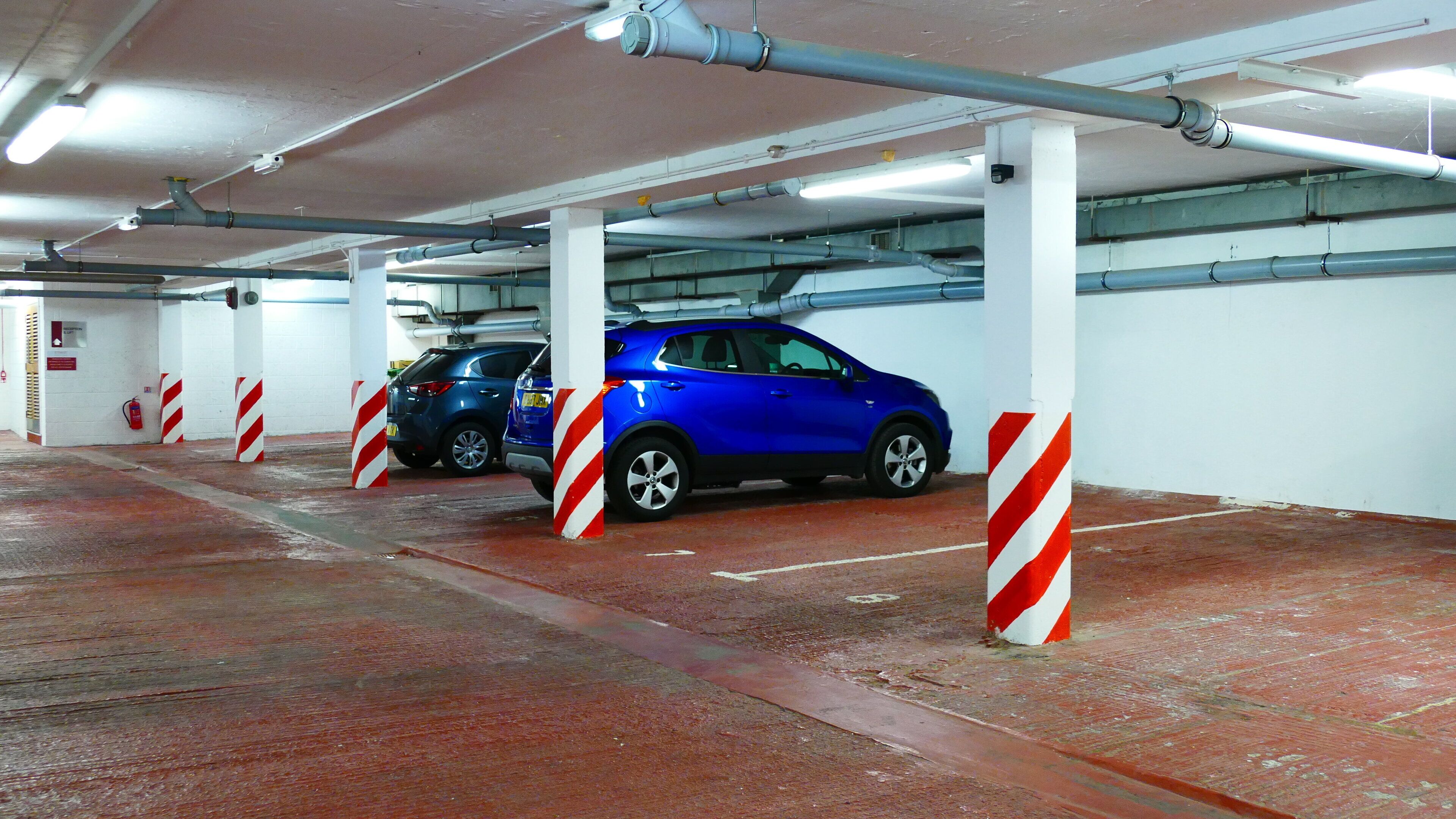 A parking garage with two cars parked under white pillars with red stripes, fluorescent lighting, and visible pipes on the ceiling.