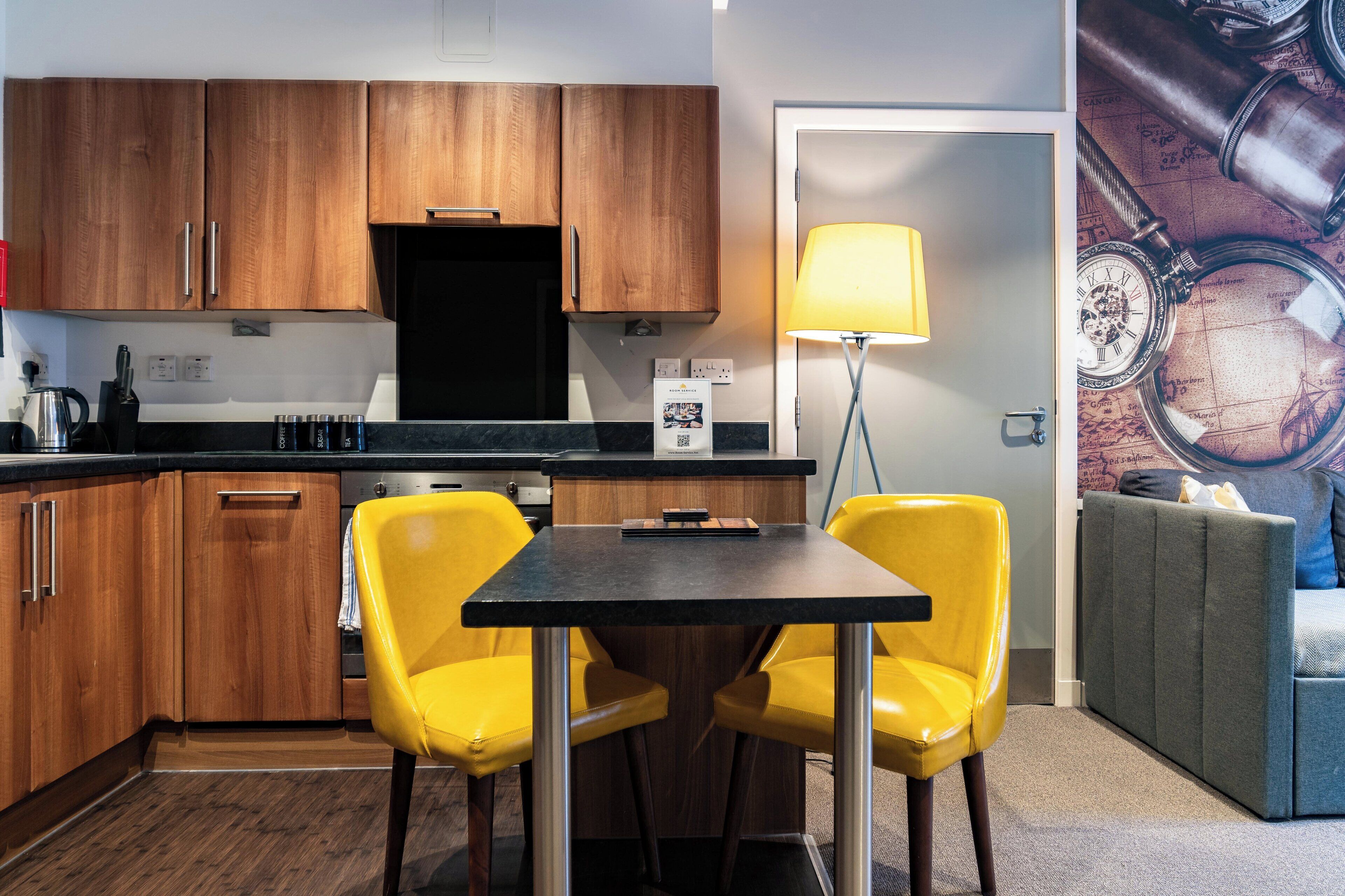 A modern kitchen with wooden cabinets, a black countertop, and a yellow dining table with two chairs.