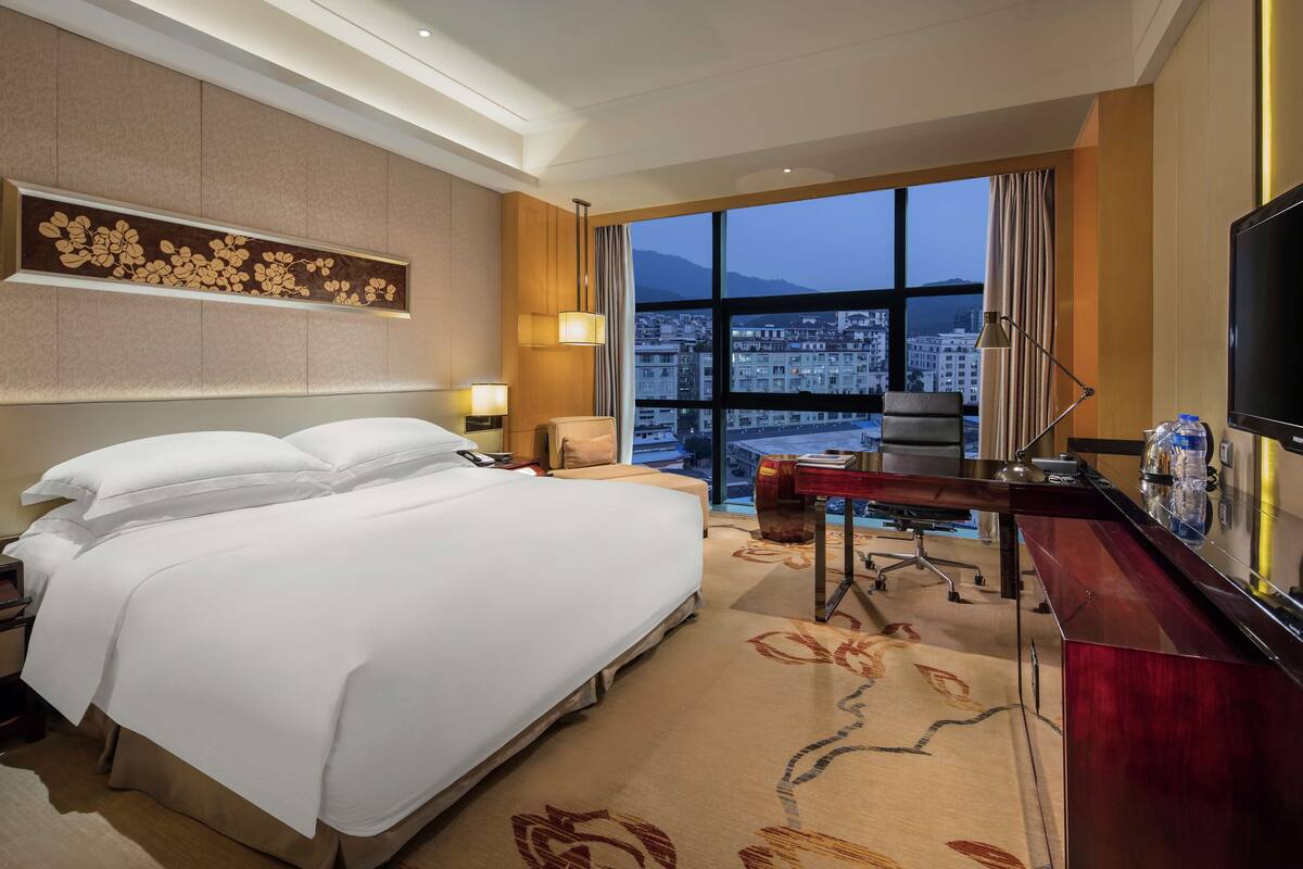 A hotel room with a large bed, a desk, a chair, and a TV. There is a window with a city view.