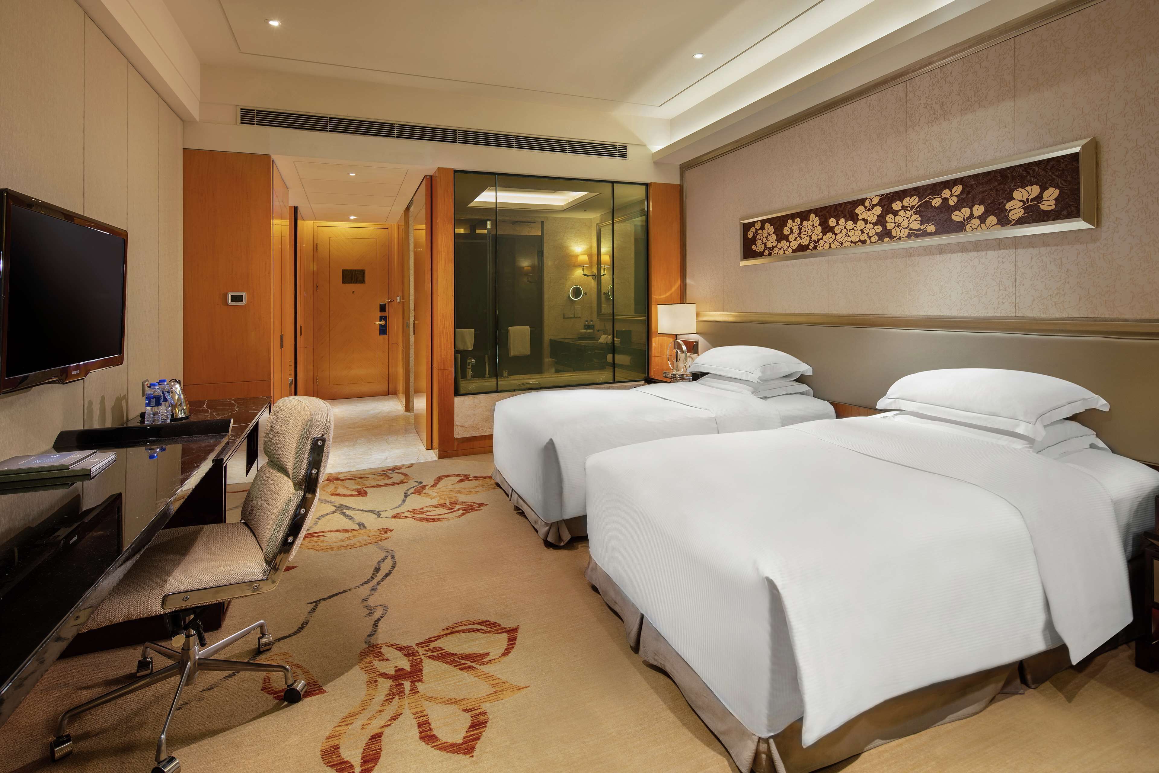 A hotel room with two beds, a desk with a chair, a television, and a bathroom visible through a glass door.