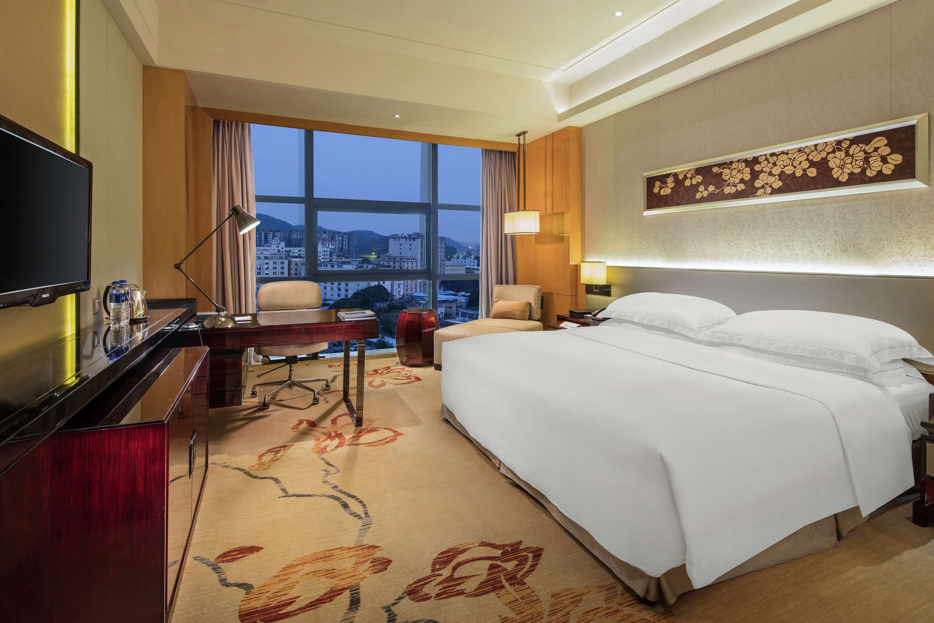 A hotel room with a large bed, a desk with a computer, a TV, and a view of the cityscape through a large window.