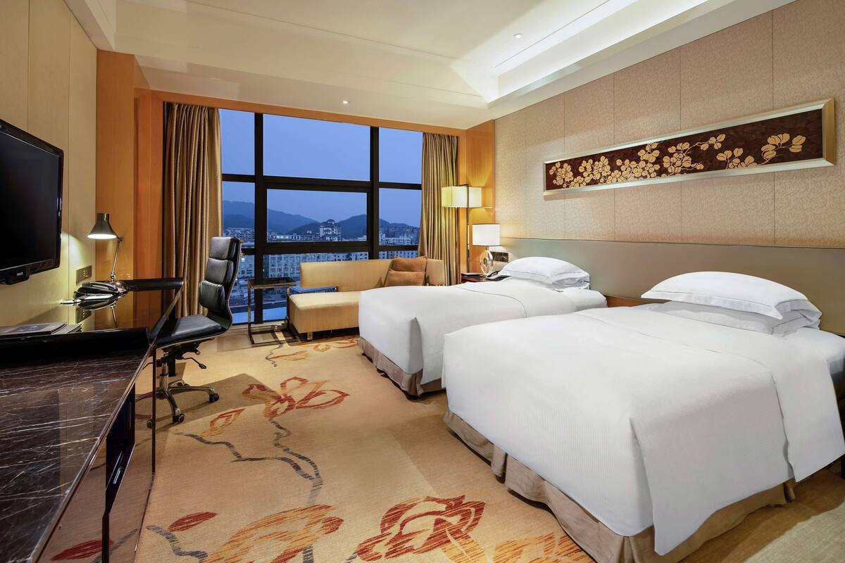A hotel room with two beds, a desk, a chair, a TV, and a large window with a city view.