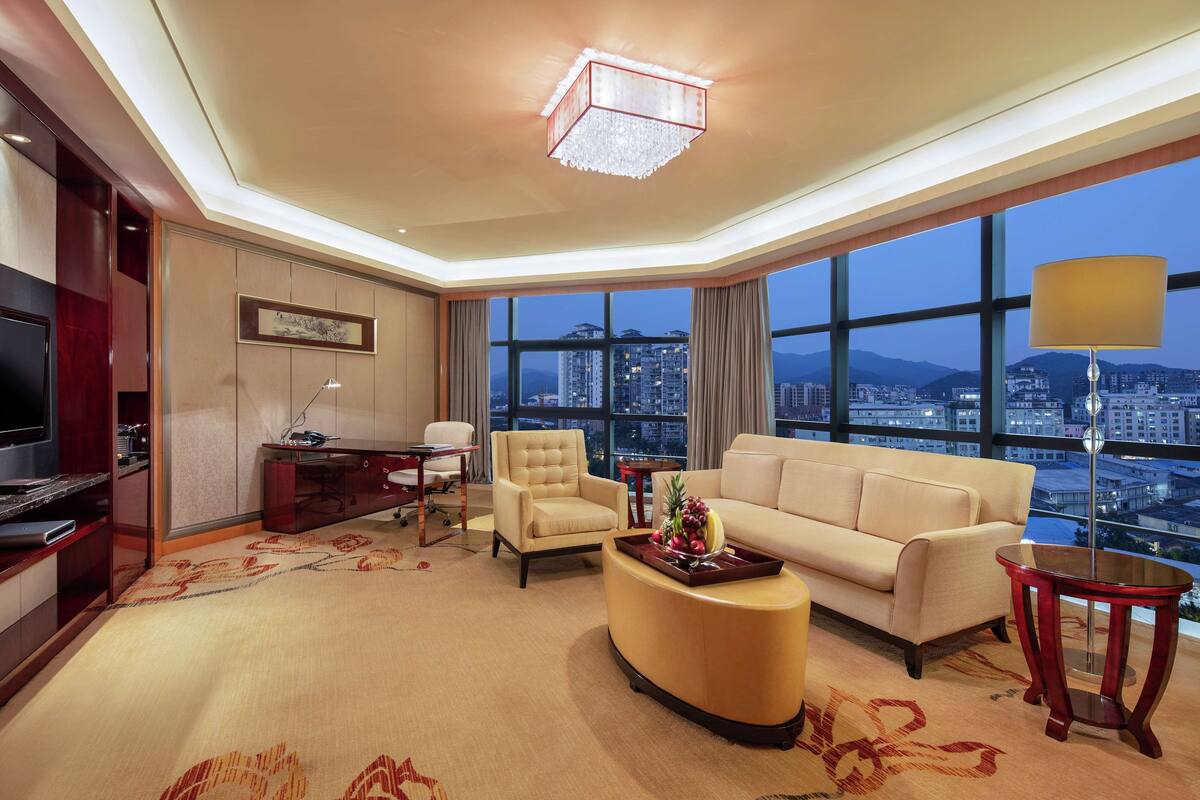 A modern hotel room with a large window offering a city view, a comfortable sofa, a desk with a lamp, and a television.