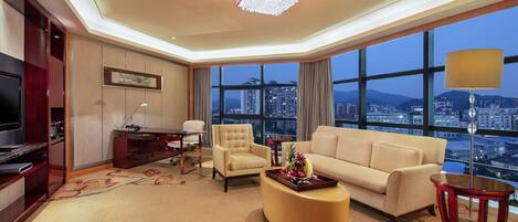 A modern hotel room with a large window offering a city view, a comfortable sofa, a desk with a lamp, and a television.