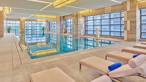 Una piscina interior moderna con paredes de vidrio transparente, sillas de descanso y un techo bien iluminado.