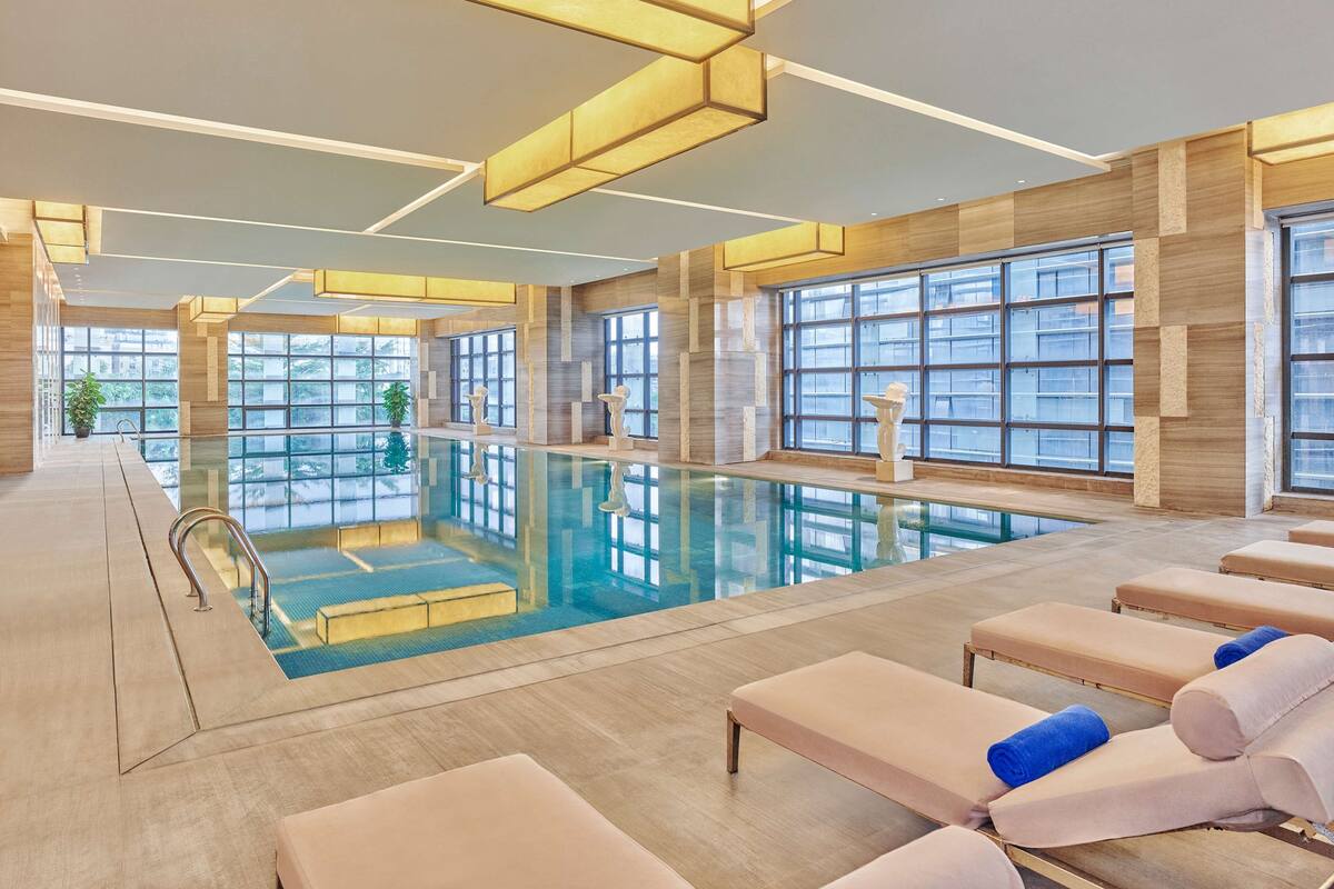 A modern indoor swimming pool with clear glass walls, lounge chairs, and a well-lit ceiling.