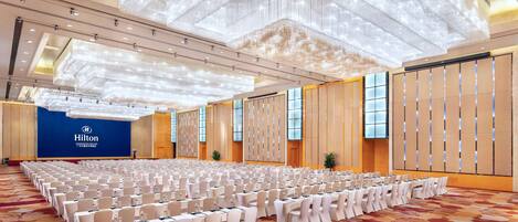 A large conference room with rows of tables and chairs, a blue wall with the Hilton logo, and a patterned carpet.