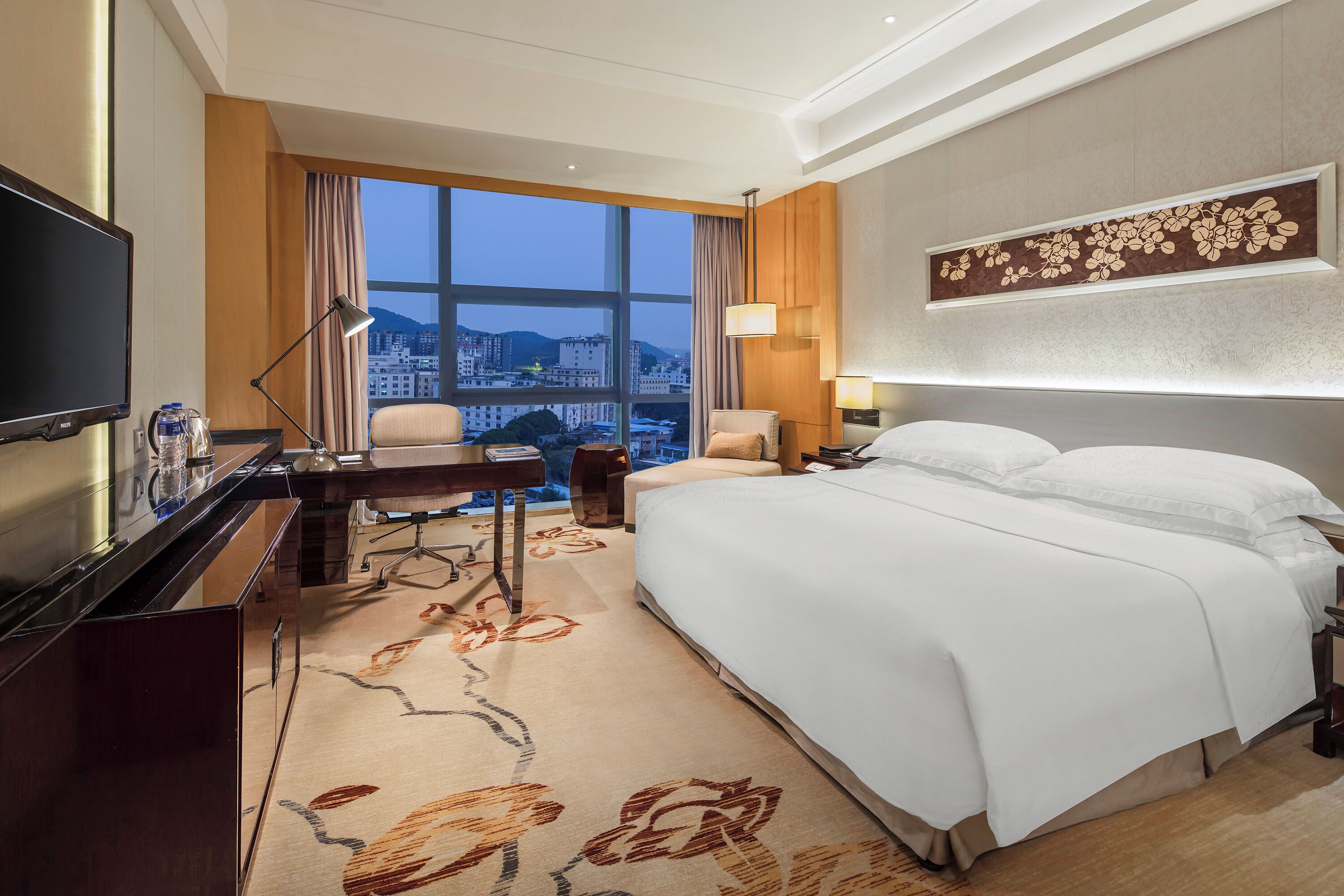 A hotel room with a large bed, a desk with a computer, a television, and a view of the cityscape through the window.