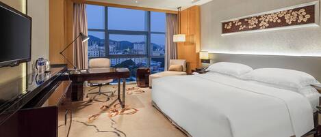 A hotel room with a large bed, a desk with a computer, a television, and a view of the cityscape through the window.
