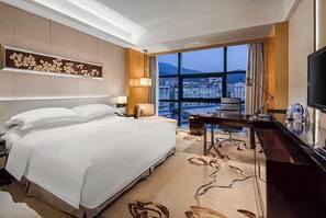 Habitación de hotel con una cama grande, un escritorio, un televisor y vistas a un paisaje urbano a través de amplias ventanas.
